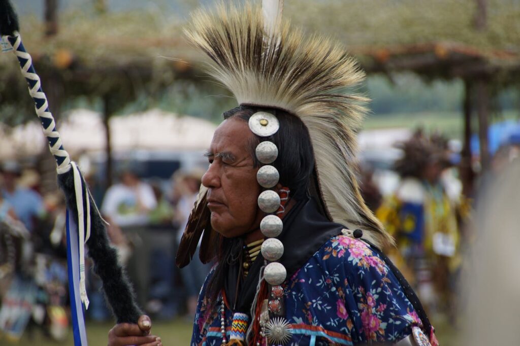An Indigenous man in traditional regalia stands in profile at an outdoor gathering. He wears a feathered headdress styled in a tall fan shape, metal conchos along the side of his head, and a floral-patterned shirt. He holds a staff wrapped with fur and blue-and-white ribbons. Other dancers and spectators are softly blurred in the background.