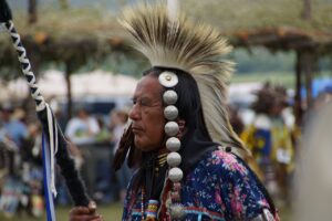 An Indigenous man in traditional regalia stands in profile at an outdoor gathering. He wears a feathered headdress styled in a tall fan shape, metal conchos along the side of his head, and a floral-patterned shirt. He holds a staff wrapped with fur and blue-and-white ribbons. Other dancers and spectators are softly blurred in the background.