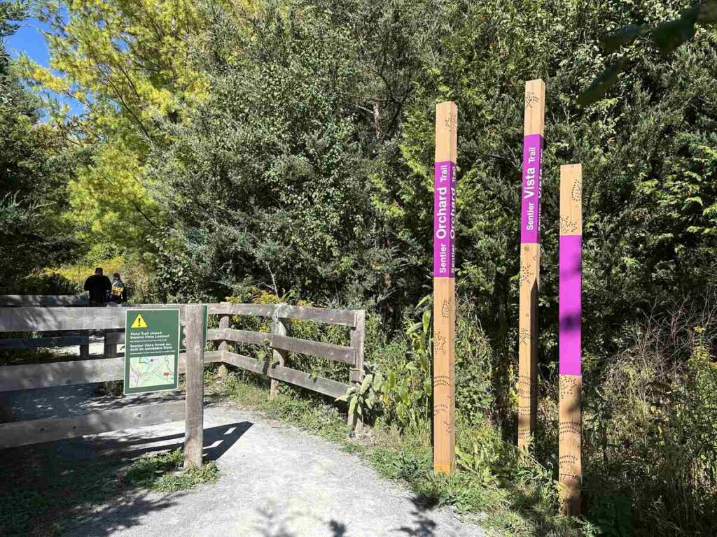A sunny trail entrance marked with Orchard Trail and Vista Trail signage, surrounded by greenery—an easy nature reset that fits beautifully into a mindful, close-to-home getaway.