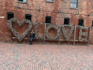 Large “LOVE” letters covered in padlocks in front of a red brick wall, with a person standing beside the sculpture.