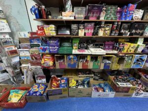 Assorted shelves and boxes filled with British crisps, chocolates and gift sets at the British Emporium, including large multi-packs of Walkers crisps stacked along the floor.