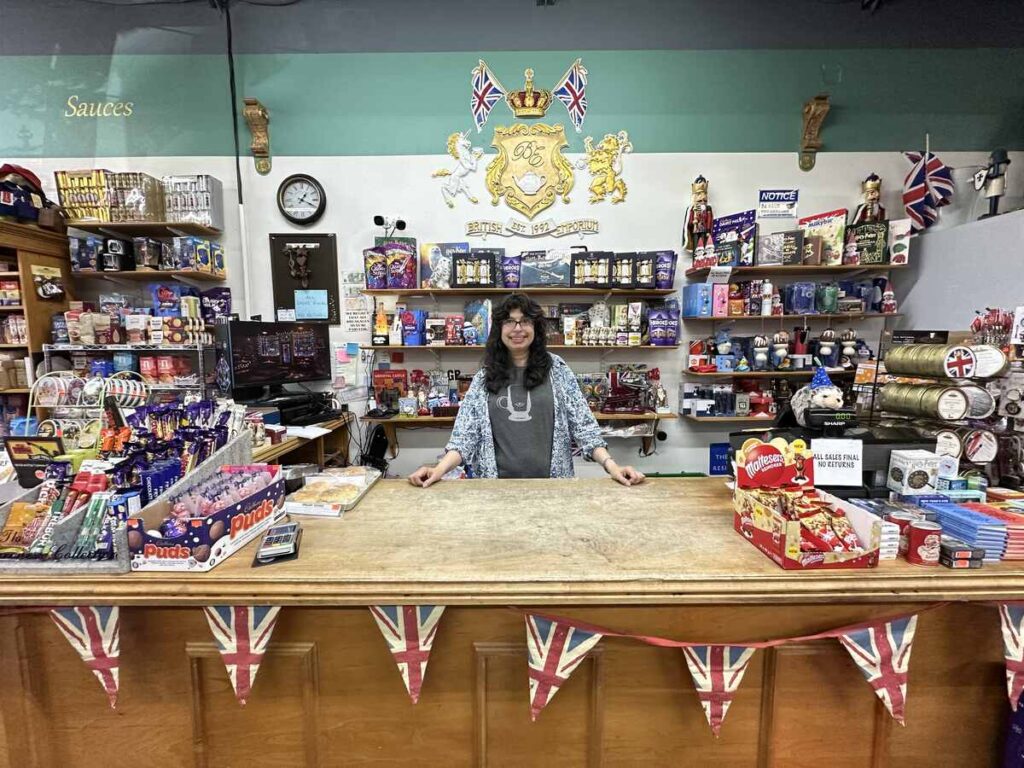 Owner Sheela Bailey standing and smiling behind the wooden counter at the British Emporium, surrounded by shelves of British chocolates, tins and souvenirs, with Union Jack bunting draped across the front.