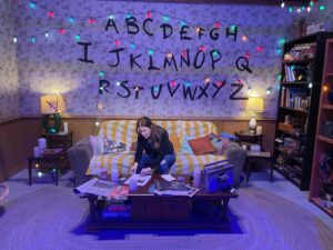 Living room set with the Stranger Things alphabet painted on the wall and colourful string lights overhead, with a person seated on a couch.