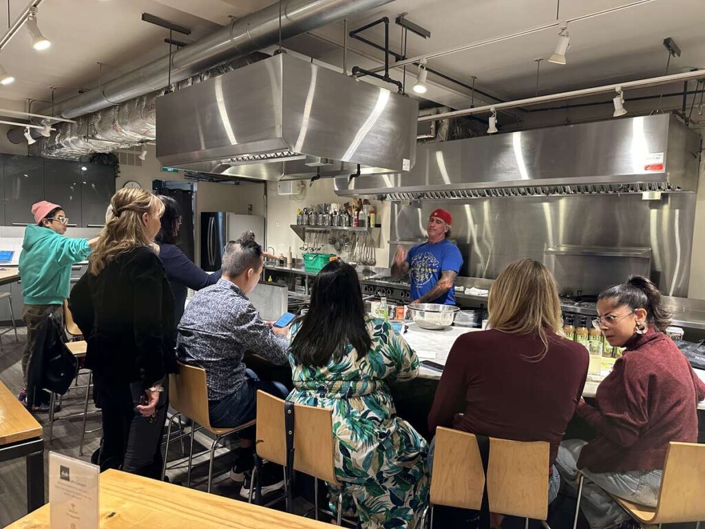 Group of people seated at a counter in a commercial kitchen while an instructor speaks from behind the work area.