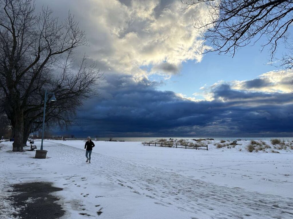 Snow-covered lakeside path with a runner in the distance, bare trees on the left, and dramatic dark clouds over the water.