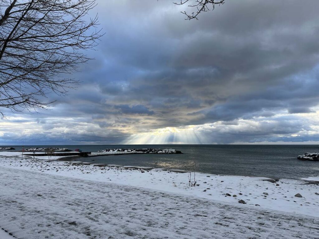Snowy lakeshore with a calm body of water, a breakwater in the distance, and sun rays streaming through dark clouds.
