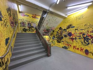 Yellow stairwell covered in graffiti and drawings with grey steps and metal handrails under bright fluorescent lights.