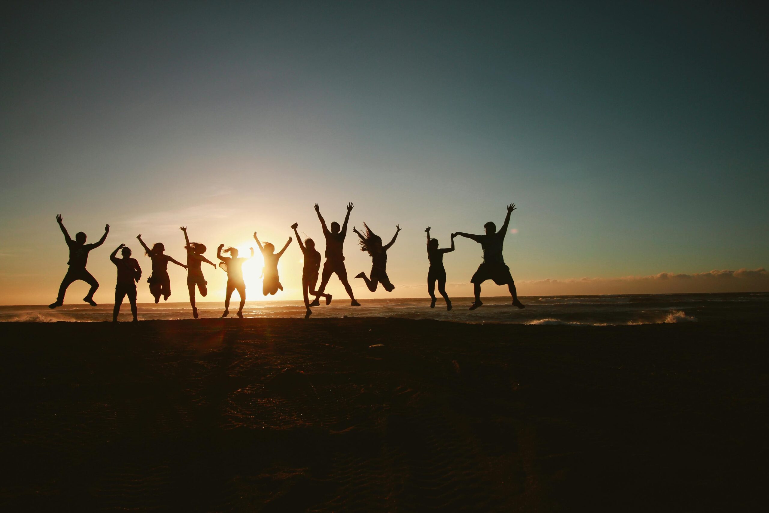 Silhouettes of a group of people jumping and cheering on a beach at sunset, with the sun low over the ocean.