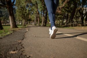 Close-up of a person’s legs in leggings and white sneakers walking or jogging along a paved park path lined with trees.