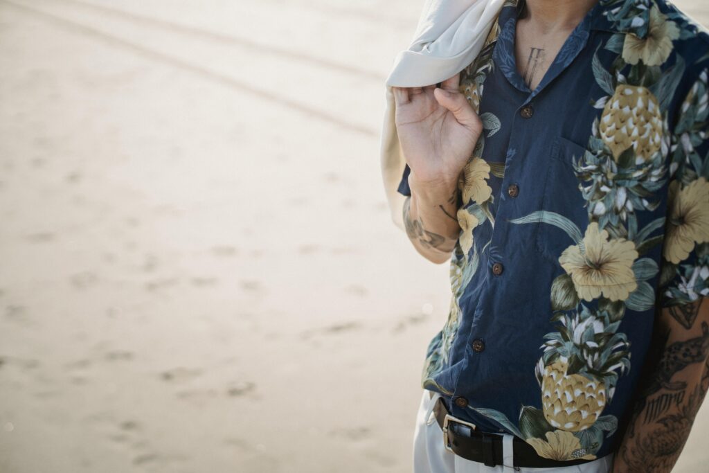 Cropped view of a person wearing a navy aloha shirt with pineapple and hibiscus prints on a sandy beach.