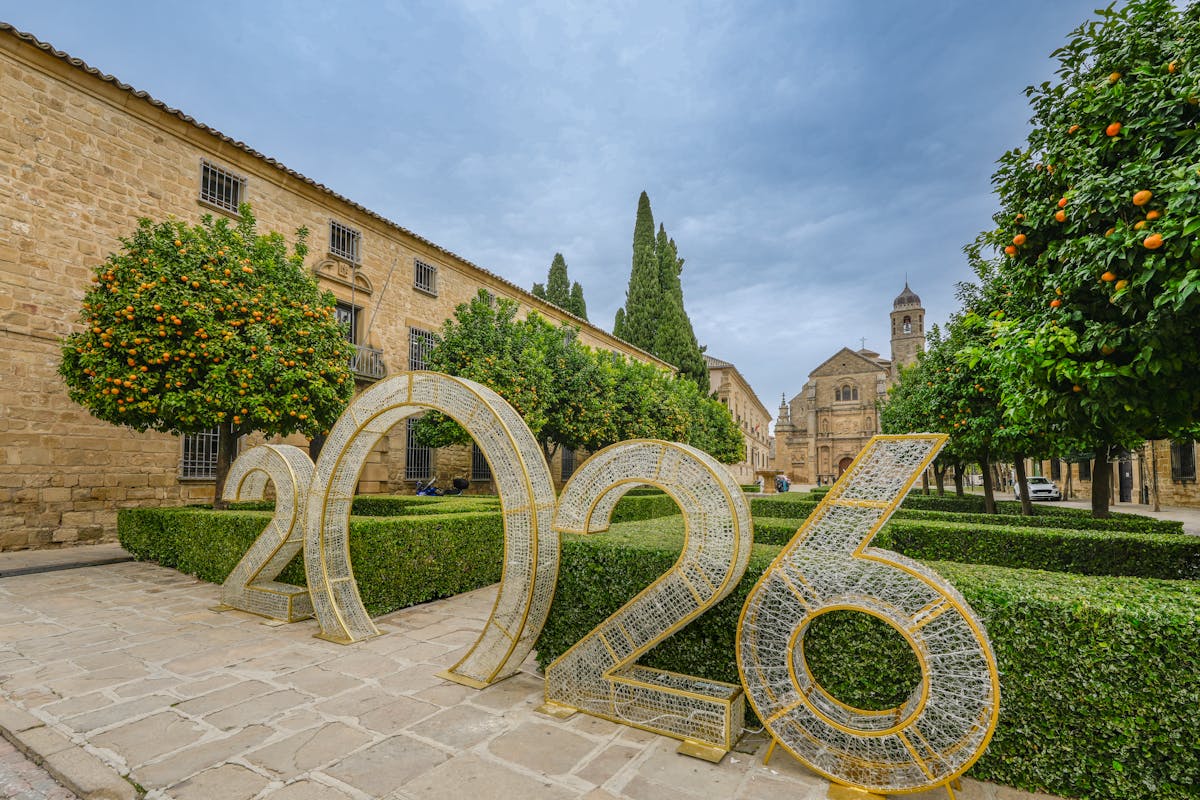 Large “2026” installation in a stone courtyard lined with orange trees, with a church in the background.