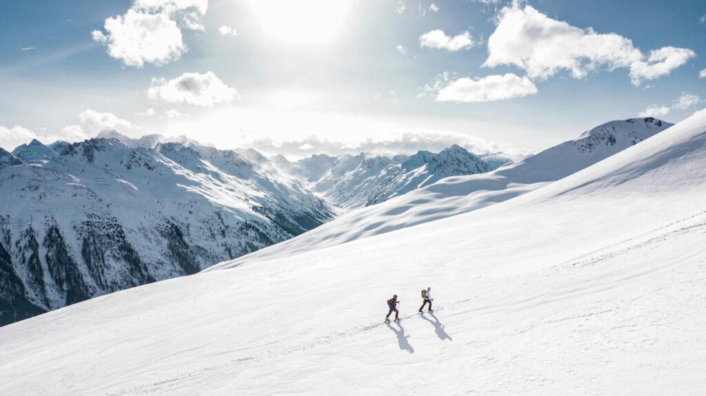 Two hikers crossing a bright snowfield with jagged mountain peaks and a low winter sun behind them.