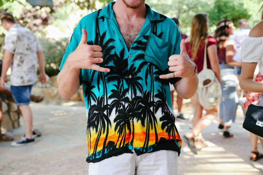Person wearing a teal aloha shirt with black palm tree silhouettes and a sunset gradient, holding both hands in the “shaka” gesture at an outdoor event.