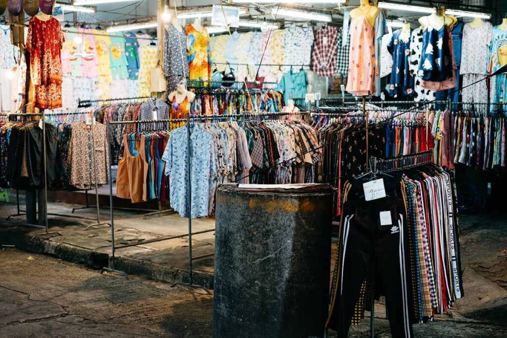 Clothing market stall with racks of patterned shirts and dresses under bright lights.