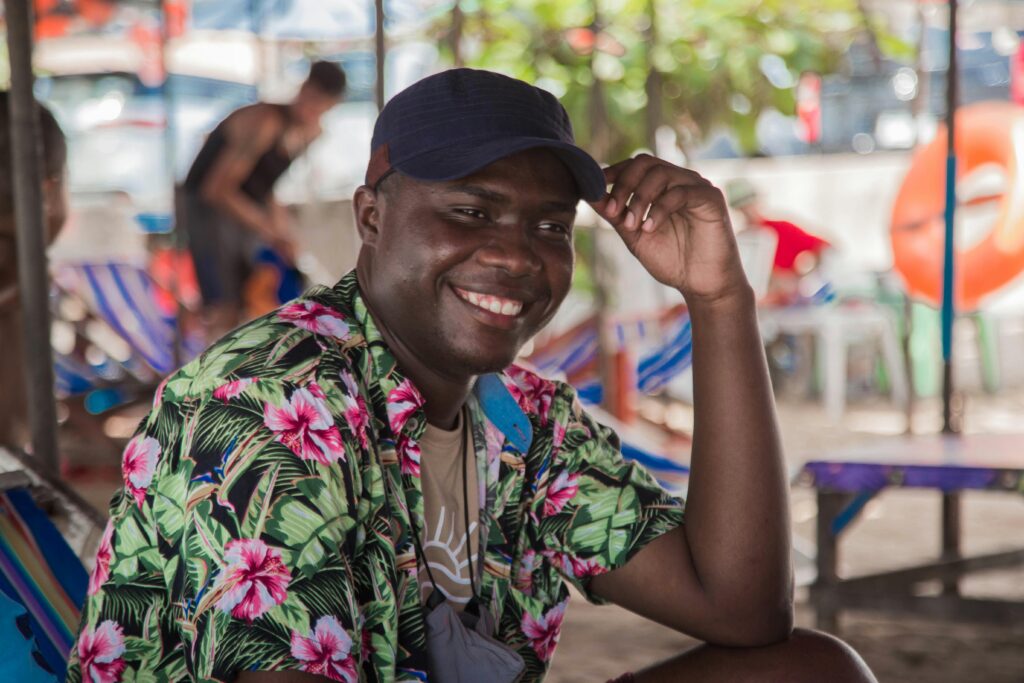 Smiling person in a green-and-pink floral aloha shirt and navy cap, seated at an outdoor beachside café.
