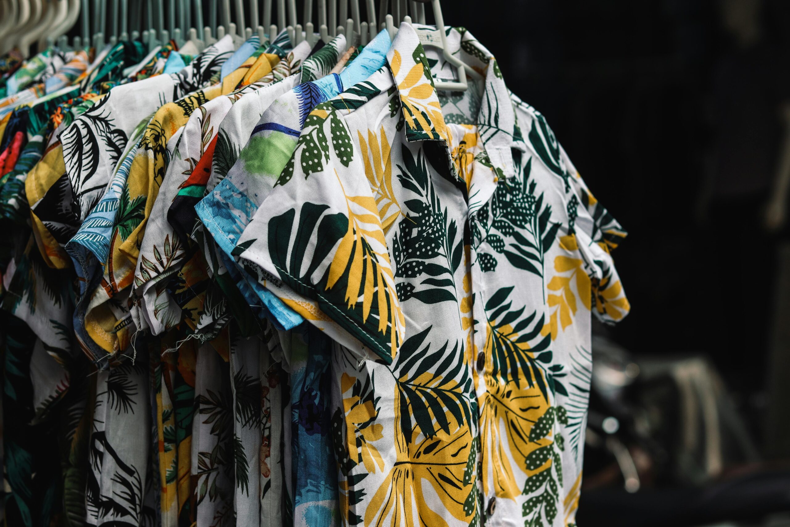 Close-up of tropical-print shirts on hangers, with palm and leaf patterns in green, yellow, and blue.