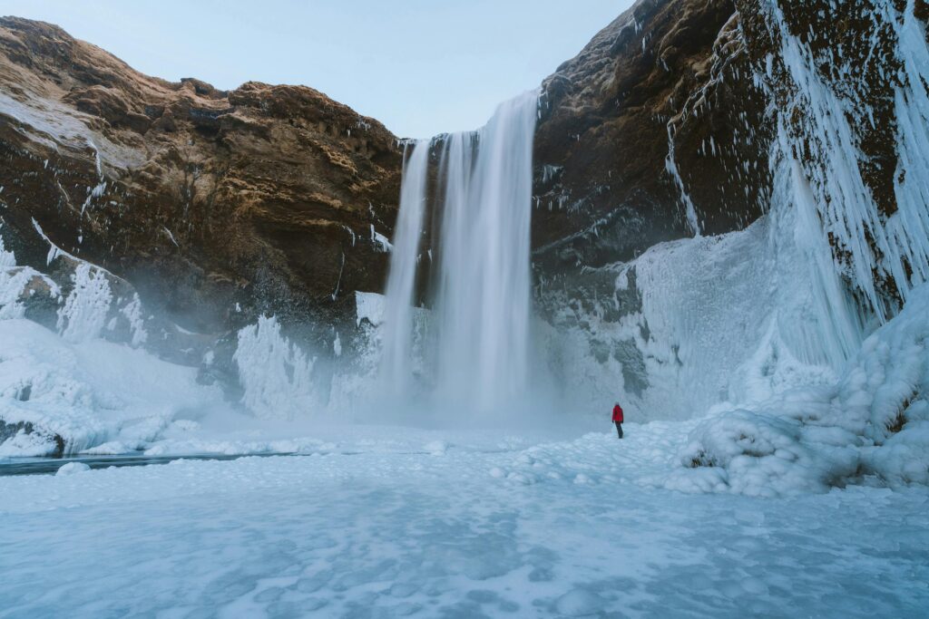 Tall waterfall pouring into an icy basin, with a person in a red jacket standing on frozen ground nearby.