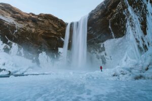 Tall waterfall pouring into an icy basin, with a person in a red jacket standing on frozen ground nearby.