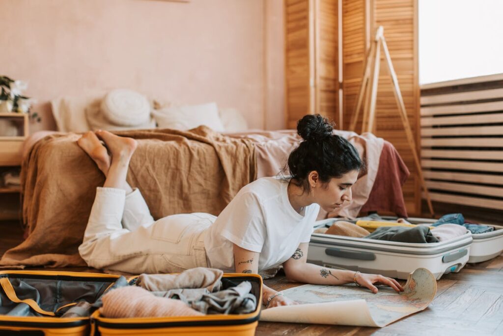 Person lying on the floor beside an open suitcase, studying a paper map in a sunlit bedroom.