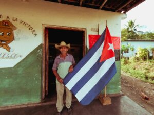 A man stands in a doorway holding a large Cuban flag outside a modest, weathered building.