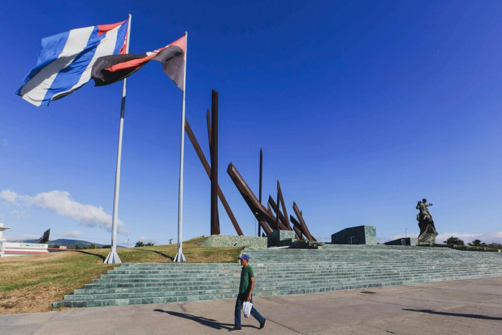 Two tall flagpoles fly large flags beside a stepped monument under a vivid blue sky; a man walks across the plaza.