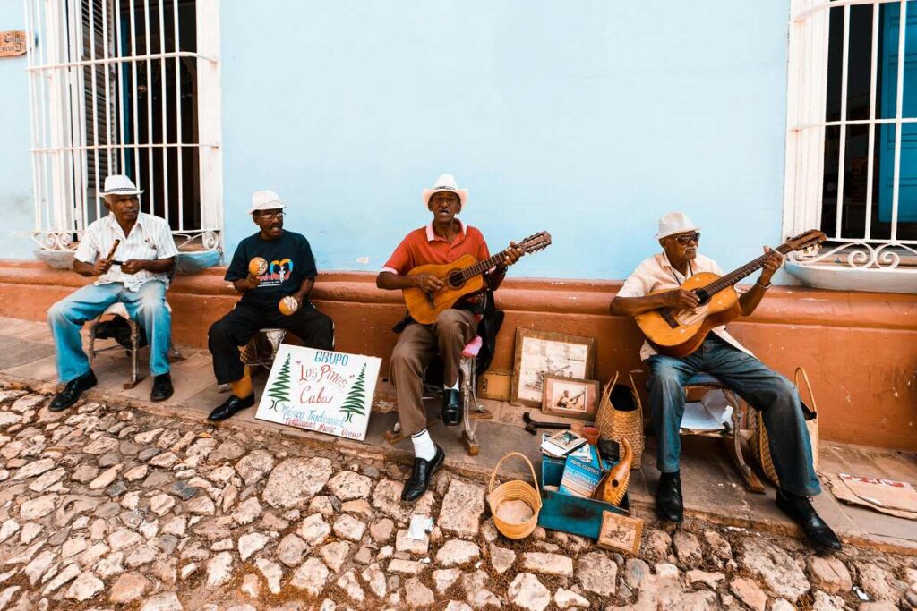 Four older men play music while seated along a pastel-blue wall on a cobblestone street; two hold guitars and one shakes maracas.