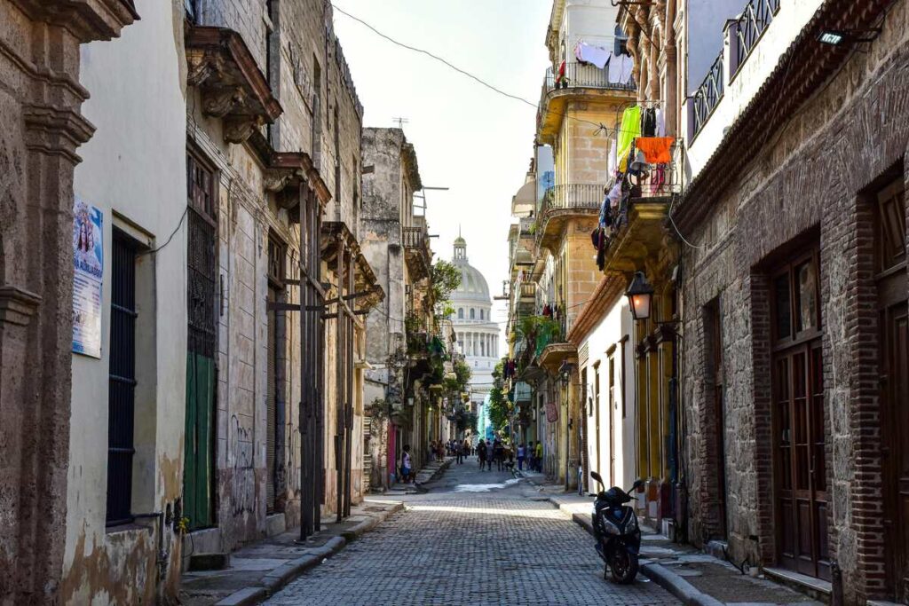 A narrow, cobblestoned street lined with worn buildings and balconies; laundry hangs above pedestrians in the distance.