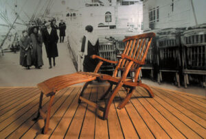 Wooden Titanic deck chair displayed on a ship-style deck floor in front of a black-and-white background image of passengers walking onboard.