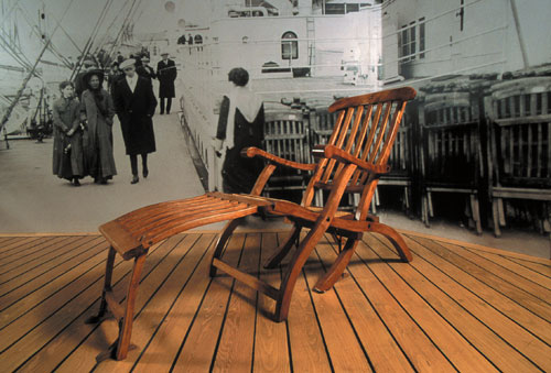 Wooden Titanic deck chair displayed on a ship-style deck floor in front of a black-and-white background image of passengers walking onboard.
