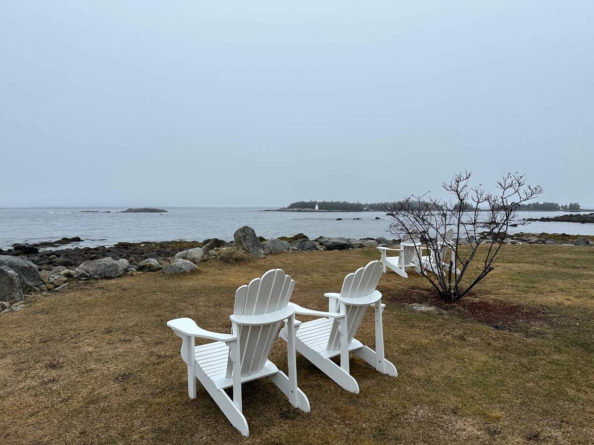 Two white Adirondack chairs face the calm, rocky shoreline at Oceanstone in Indian Harbour, Nova Scotia.