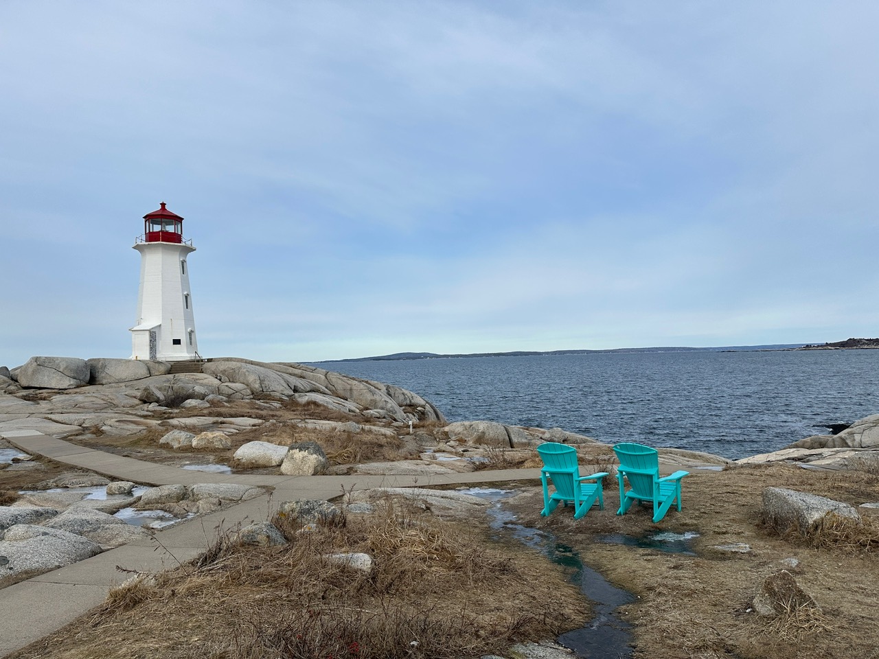 Peggys Point Lighthouse stands on pale granite beside the Atlantic Ocean, while two turquoise Adirondack chairs face the water under a soft grey-blue sky.
