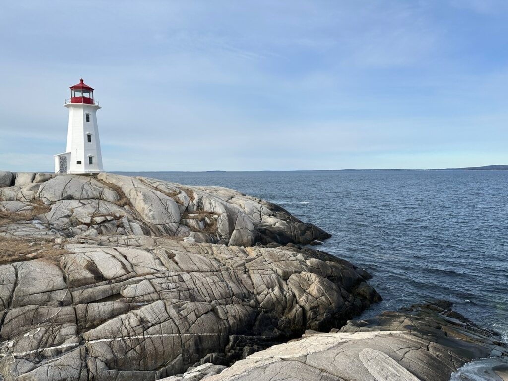 Peggys Point Lighthouse stands on smooth pale granite at Peggy’s Cove, with the Atlantic Ocean stretching across the background under a soft blue sky.