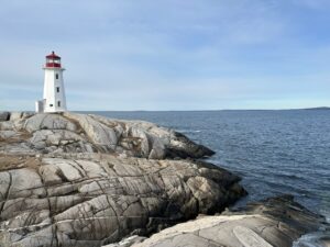 Peggys Point Lighthouse stands on smooth pale granite at Peggy’s Cove, with the Atlantic Ocean stretching across the background under a soft blue sky.