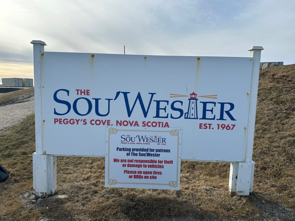 White roadside sign for The Sou’Wester in Peggy’s Cove, with a smaller parking notice mounted below it.