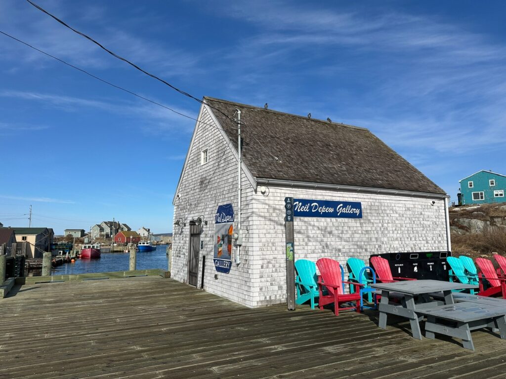 Neil Depew Gallery stands on the Peggy’s Cove waterfront beside bright red and turquoise Adirondack chairs, with fishing boats and houses in the harbour behind it.