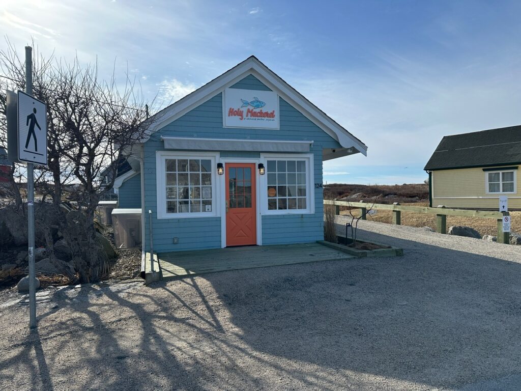 Small pale blue shop with an orange door and a sign reading Holy Mackerel sits beside a gravel path in Peggy’s Cove.