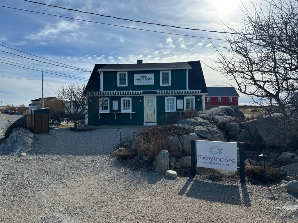 Dark teal building with a sign reading Hunky Dory stands behind rocks and gravel in Peggy’s Cove, with a sign for Six by the Sea in the foreground.