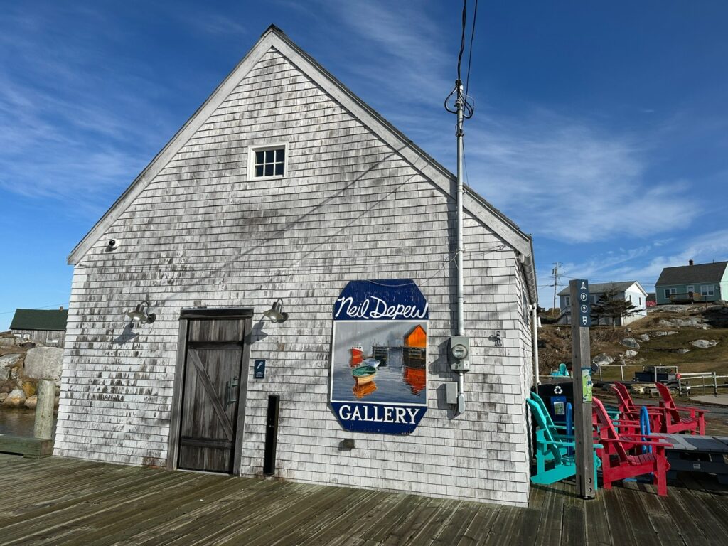 White shingled building marked Neil Depew Gallery stands beside the boardwalk and colourful outdoor chairs in Peggy’s Cove.