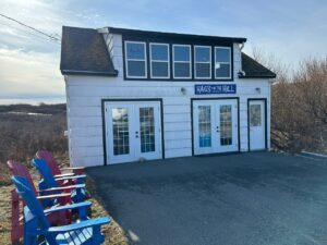 Small white building with a sign reading Hags on the Hill, with two brightly coloured Adirondack chairs at the side and low brush behind it.