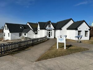 White deGarthe Gallery building with black trim and a sign at the entrance, seen from the sidewalk on a clear day.