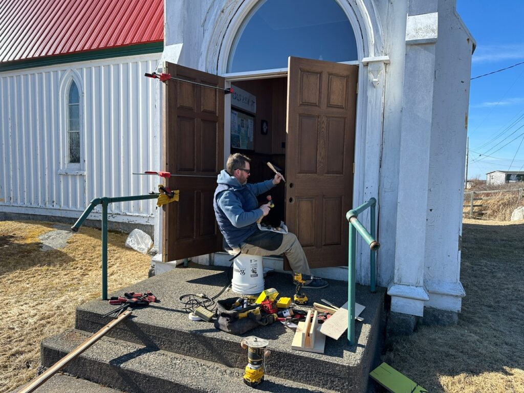 A craftsman sits on the front steps of St. John’s Anglican Church in Peggy’s Cove, repairing one of the large wooden entrance doors with tools spread around him.