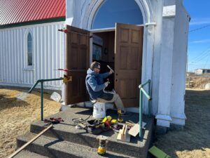 A craftsman sits on the front steps of St. John’s Anglican Church in Peggy’s Cove, repairing one of the large wooden entrance doors with tools spread around him.