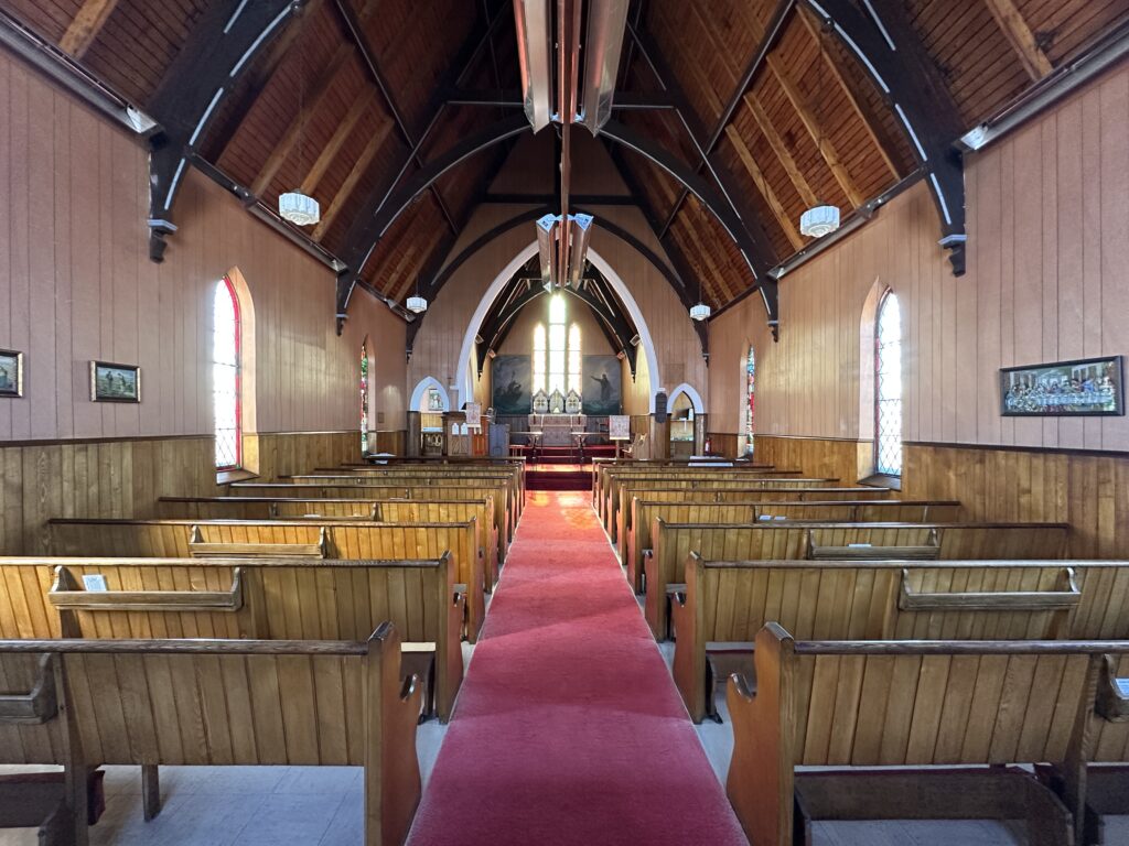 Interior of St. John’s Anglican Church in Peggy’s Cove, showing wooden pews, a red carpet aisle, tall arched windows, and a timber roof overhead.