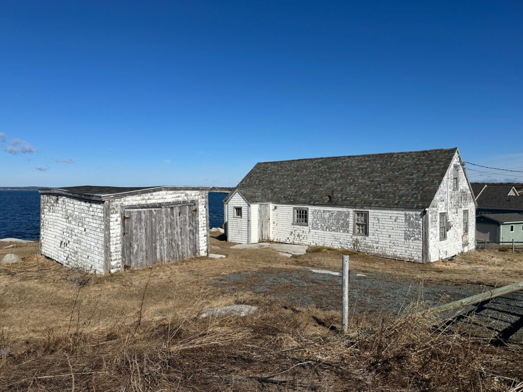A weathered white house and separate outbuilding stand on dry grass beside the ocean in Peggy’s Cove under a bright blue sky.