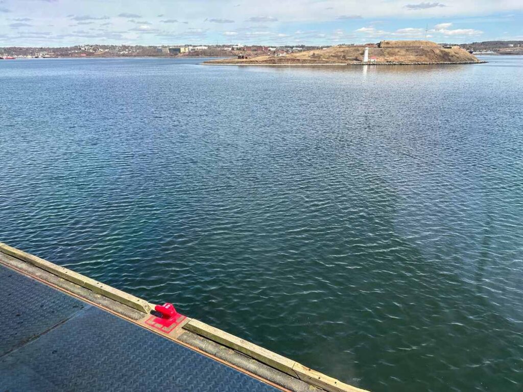 View of Halifax Harbour from Pier 21, with calm blue water in the foreground and Georges Island visible in the distance. Red pier 21 marker.