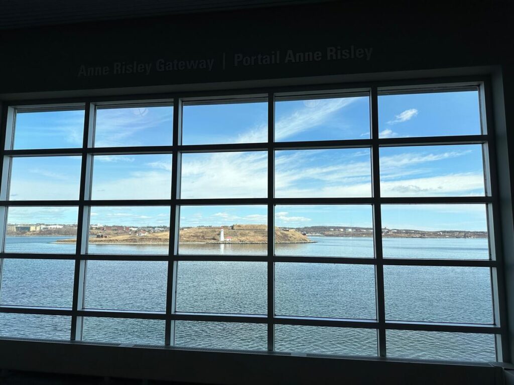 Large grid window inside Pier 21 framing a bright view of Halifax Harbour and Georges Island beyond the glass.