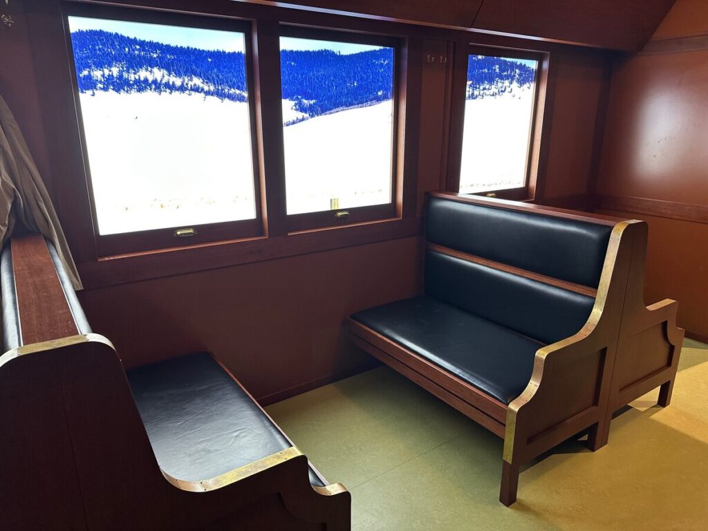 Interior of a recreated colonist train car with wooden walls, bench seating, and bright windows displaying a snowy landscape scene.