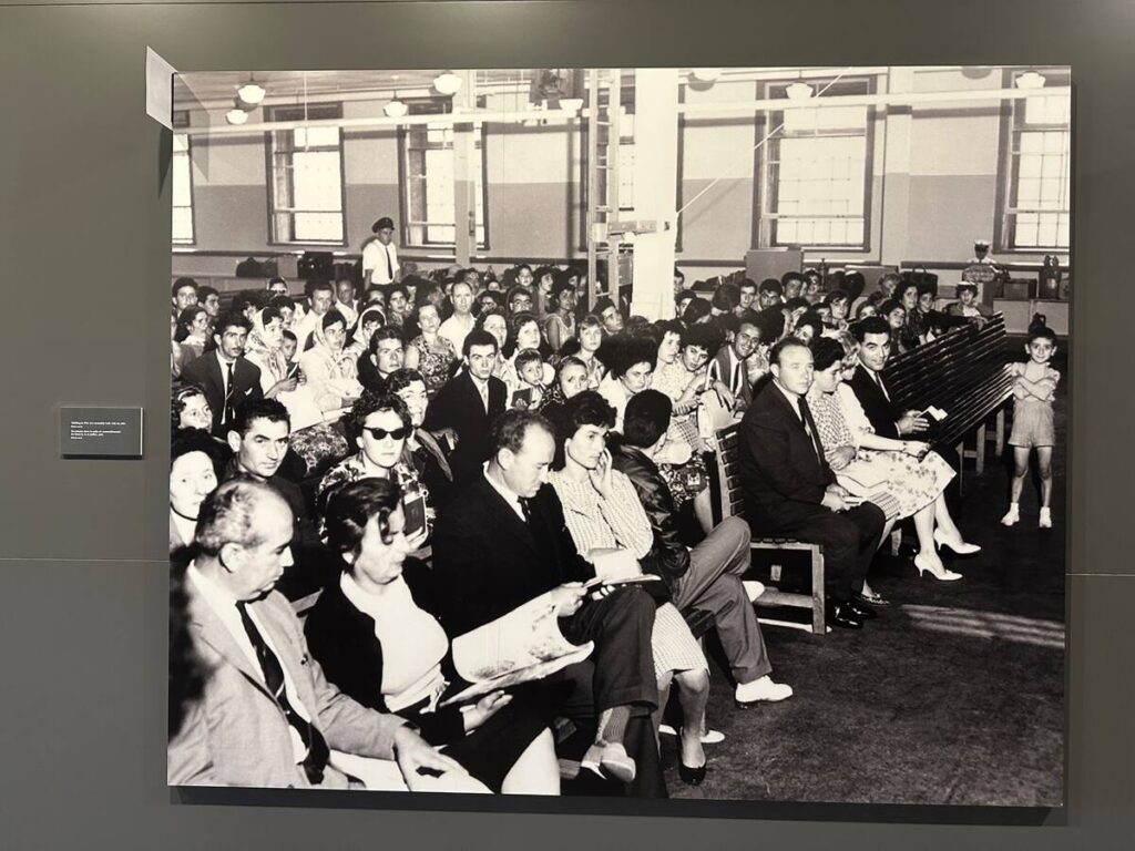 Large black-and-white photograph in the museum showing immigrants seated closely together in an assembly hall while waiting to be processed.