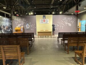 Recreated Pier 21 processing hall with rows of wooden benches facing a desk beneath a multilingual “Welcome to Canada” sign.