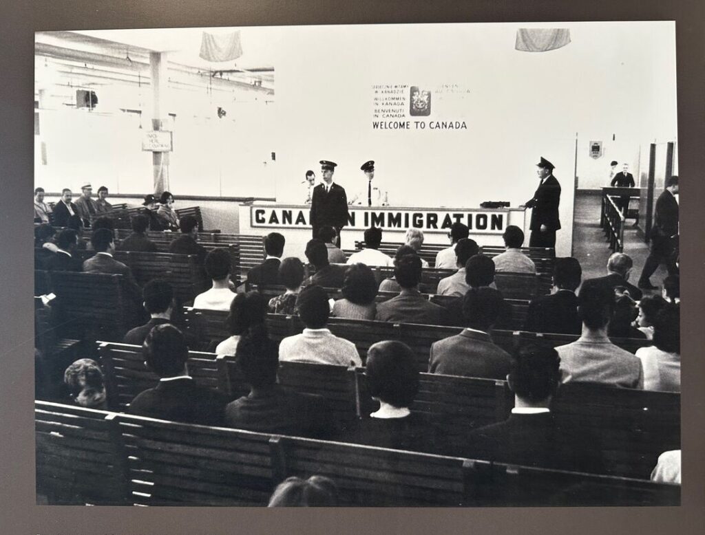 Black-and-white museum photograph showing immigrants seated on wooden benches in a processing hall beneath a “Welcome to Canada” sign while officials stand at the front.